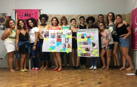Treze mulheres posam com dois cartazes em um ambiente de sala de aula.