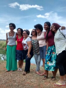 Fotografia de sete mulheres abraçadas olhando para o horizonte. atrás delas vê-se o céu azul.