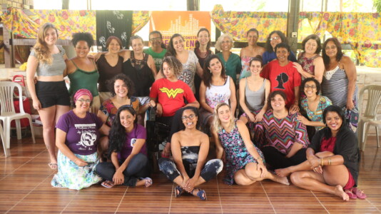 Fotografia de 25 mulheres, com fundo com alguns tecidos coloridos.
