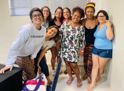 Fotografia de 8 mulheres abraçadas em um ambiente de sala de aula.