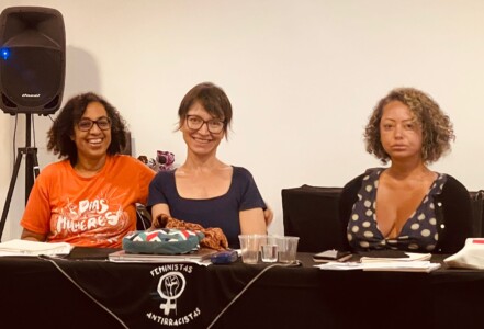 Foto de três mulheres sentadas atrás de uma mesa com toalha preta.